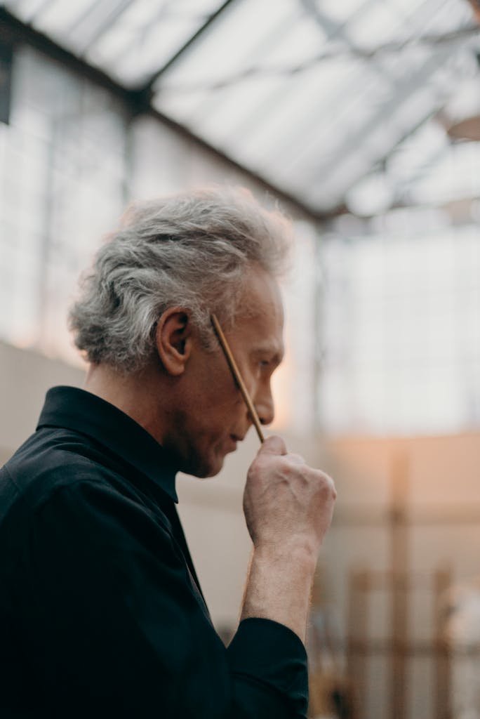 Senior artist pondering with a paintbrush in a well-lit studio during daytime.